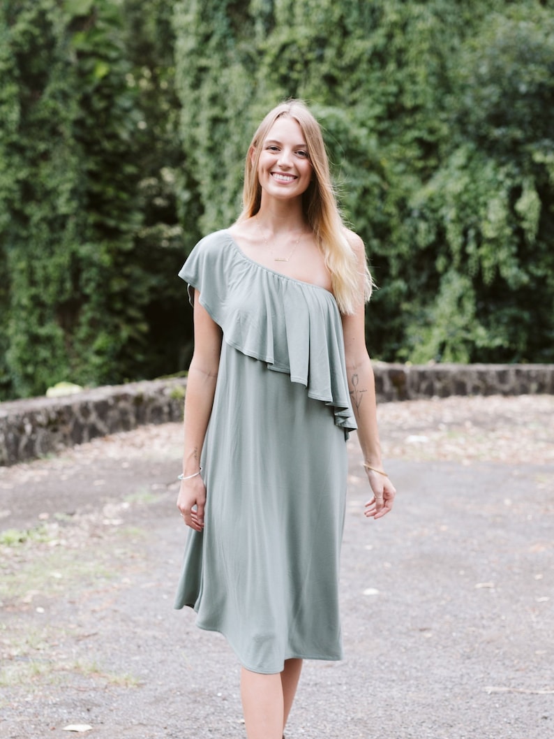 May include: A woman wearing a sage green one-shoulder dress with a ruffle detail. She is standing in front of a green leafy background.