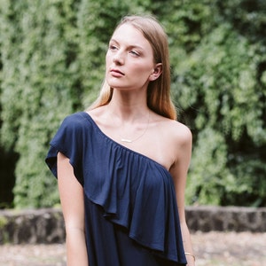 May include: A woman wearing a one-shoulder navy blue dress with a ruffled neckline. She is standing in front of a green leafy background.