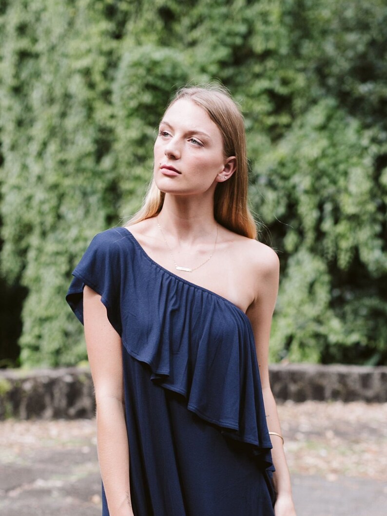 May include: A woman wearing a one-shoulder navy blue dress with a ruffled neckline. She is standing in front of a green leafy background.