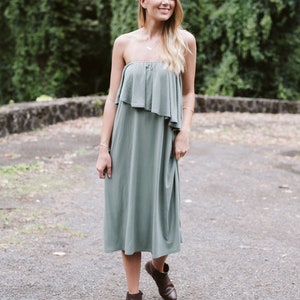 May include: A woman wearing a sage green strapless dress with a ruffled top and brown ankle boots. She is standing on a gravel path with green foliage in the background.