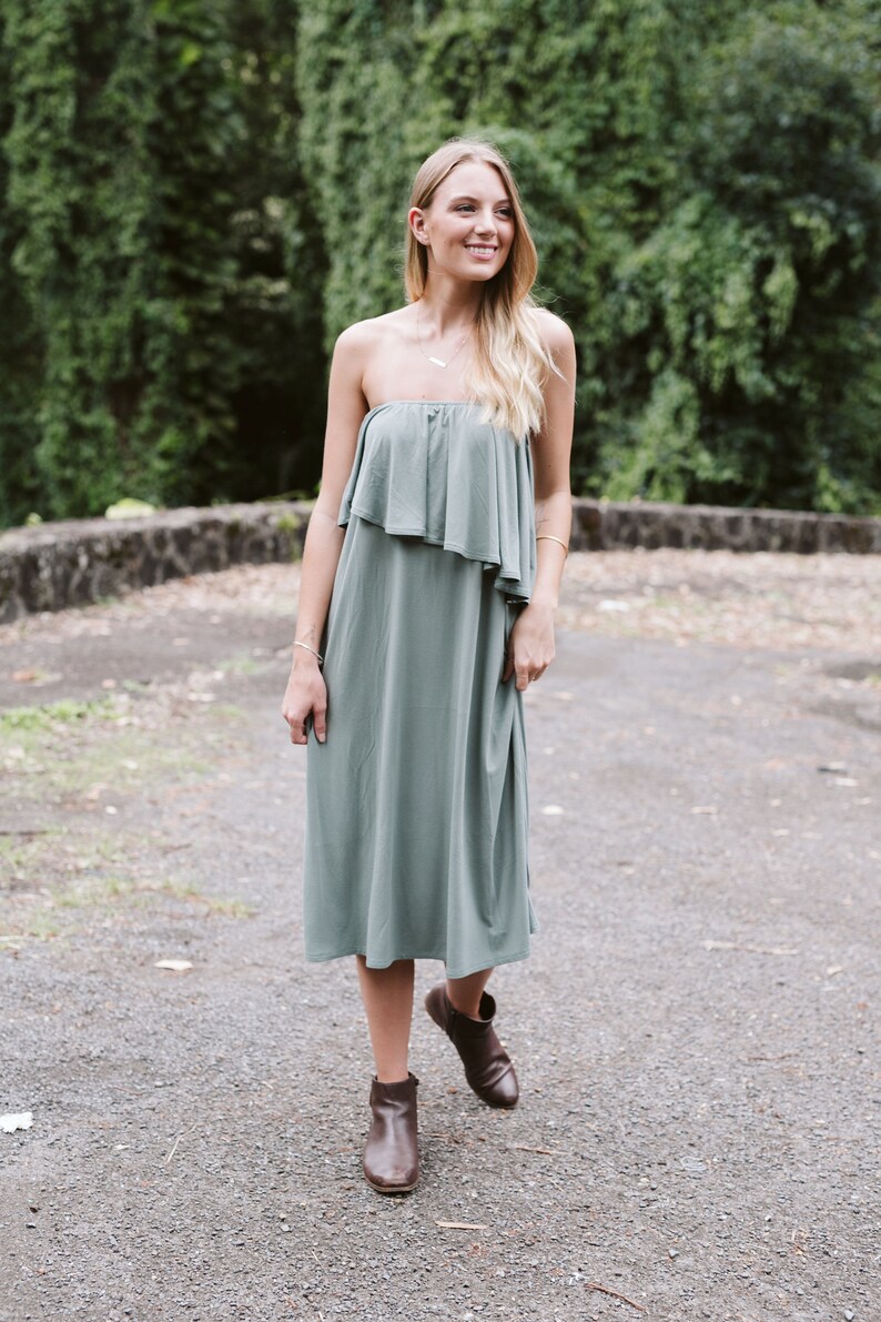 May include: A woman wearing a sage green strapless dress with a ruffled top and brown ankle boots. She is standing on a gravel path with green foliage in the background.
