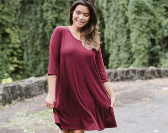 May include: A woman wearing a maroon, 3/4 sleeve, knee-length, loose-fitting dress with a scoop neck.