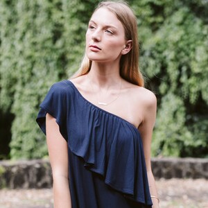May include: A woman wearing a one-shoulder navy blue dress with a ruffle detail. She is standing in front of a green leafy background.