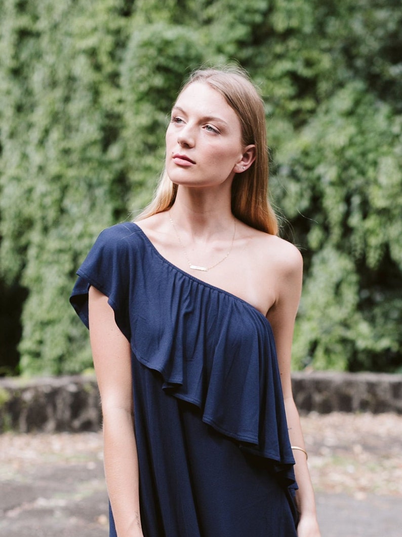 May include: A woman wearing a one-shoulder navy blue dress with a ruffle detail. She is standing in front of a green leafy background.