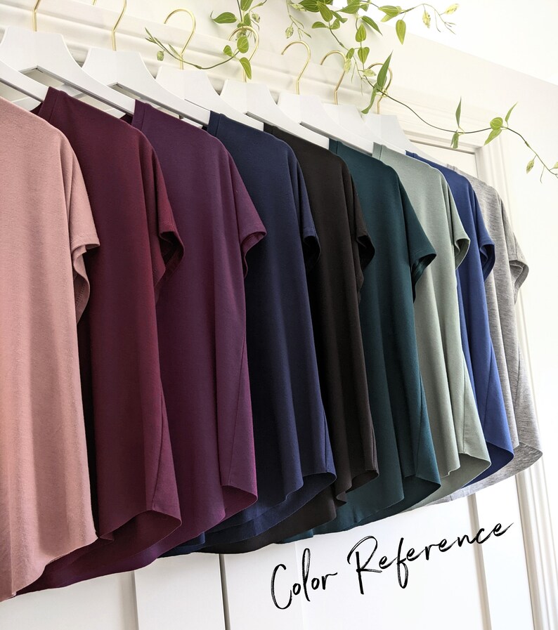 May include: A row of ten t-shirts hanging on a white rack, showcasing a variety of colors including dusty rose, burgundy, navy blue, black, teal, sage green, and gray. The text "Color Reference" is written in cursive at the bottom of the image.