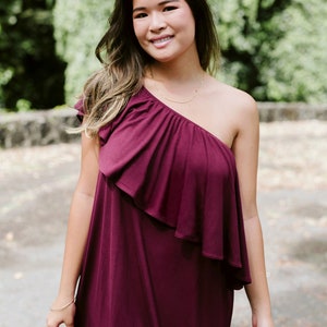 May include: A woman wearing a one-shoulder burgundy dress with a ruffled neckline. She is standing outdoors with green foliage in the background.