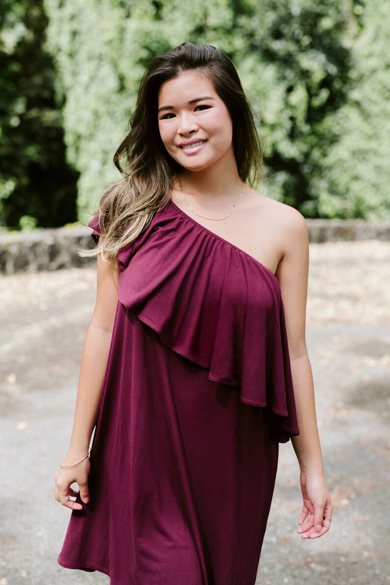May include: A woman wearing a one-shoulder burgundy dress with a ruffled neckline. She is standing outdoors with green foliage in the background.