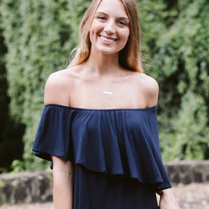 May include: A woman wearing a navy blue off-the-shoulder dress with a ruffled neckline. She is smiling and has long blonde hair.