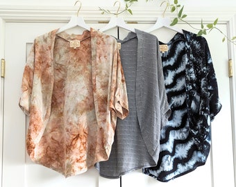 May include: Three open-front kimonos hanging on white hangers. The first is a tan and brown tie-dye pattern, the second is a grey fabric with thin white vertical stripes, and the third is a black and blue tie-dye pattern with a wavy design.