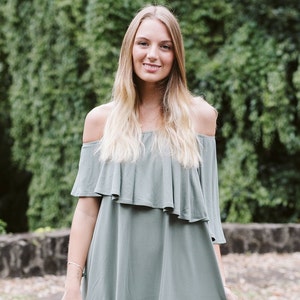 May include: A woman wearing a light green off-the-shoulder dress with a ruffled neckline. She is standing in front of a green leafy background.