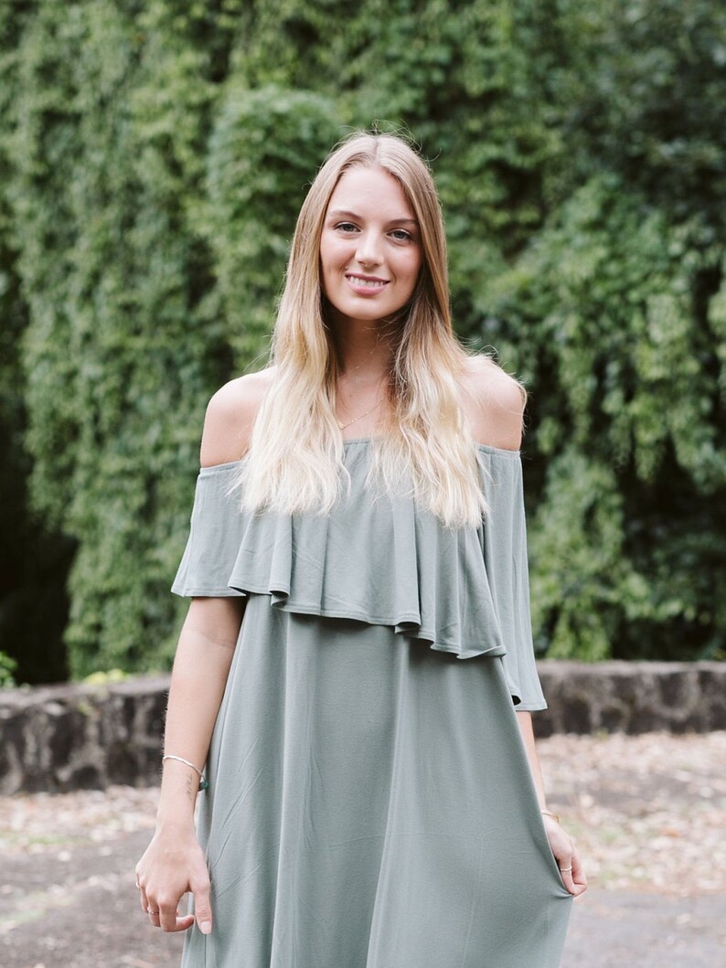May include: A woman wearing a light green off-the-shoulder dress with a ruffled neckline. She is standing in front of a green leafy background.