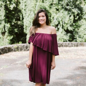 May include: A woman wearing a burgundy off-the-shoulder dress with a ruffled neckline. She is standing in front of a green leafy background.