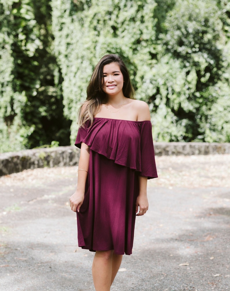 May include: A woman wearing a burgundy off-the-shoulder dress with a ruffled neckline. She is standing in front of a green leafy background.