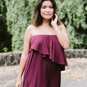 May include: A woman wearing a strapless burgundy dress with a ruffled top. She is standing in front of a green leafy background.