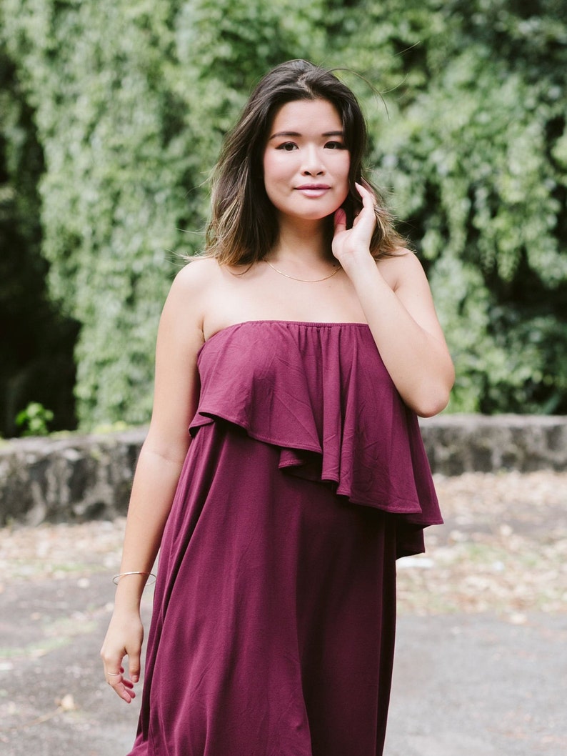 May include: A woman wearing a strapless burgundy dress with a ruffled top. She is standing in front of a green leafy background.