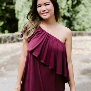 May include: A woman wearing a one-shoulder burgundy dress with a ruffled neckline. She is standing in front of a green background.