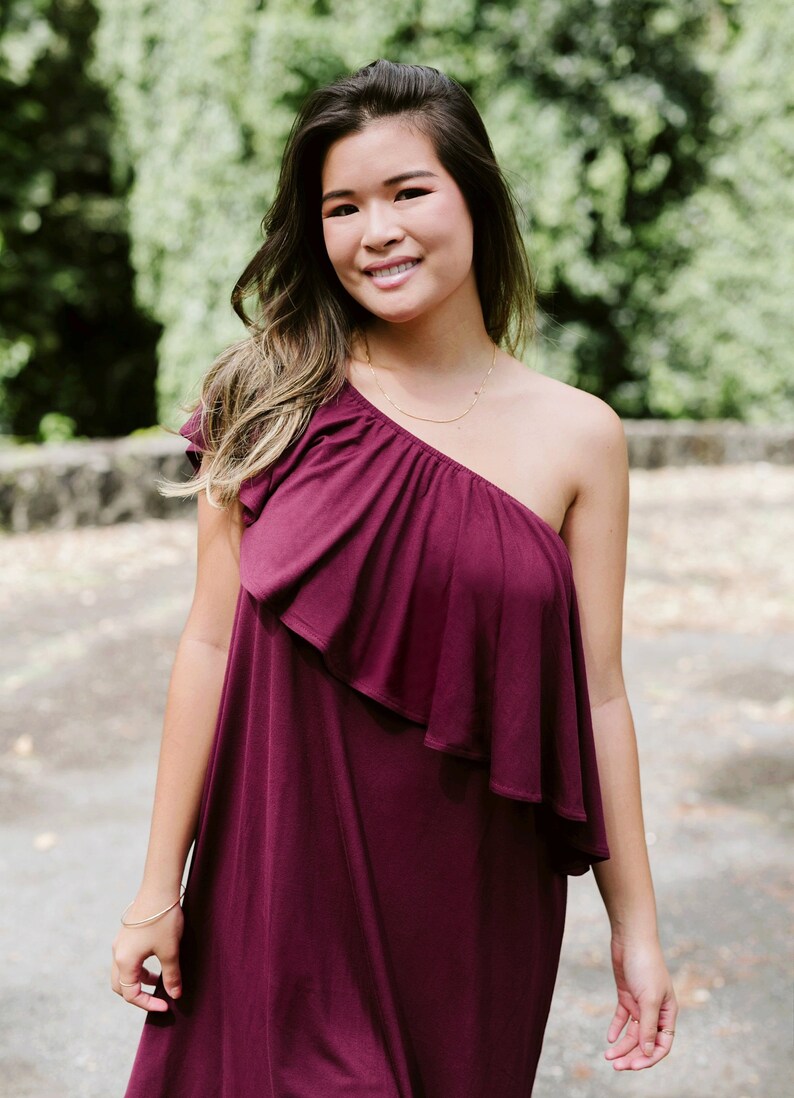 May include: A woman wearing a one-shoulder burgundy dress with a ruffled neckline. She is standing in front of a green background.