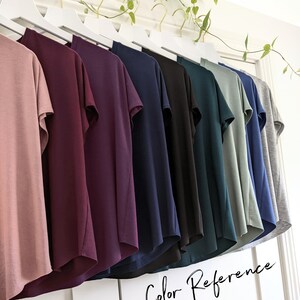 May include: A row of ten t-shirts hanging on a white rack, showcasing a variety of colors including dusty rose, burgundy, navy blue, black, teal, sage green, and gray. The text "Color Reference" is written in cursive at the bottom of the image.