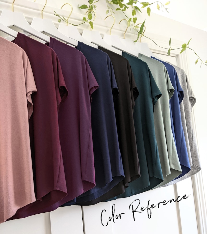 May include: A row of ten t-shirts hanging on a white rack, showcasing a variety of colors including dusty rose, burgundy, navy blue, black, teal, sage green, and gray. The text "Color Reference" is written in cursive at the bottom of the image.