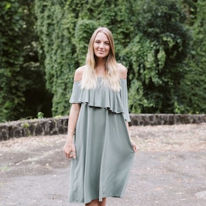 May include: A woman wearing a light green off-the-shoulder dress with a ruffle detail at the neckline. She is standing in front of a green leafy background.