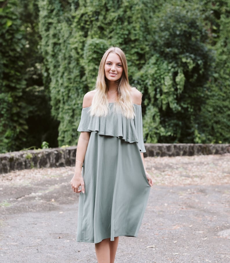 May include: A woman wearing a light green off-the-shoulder dress with a ruffle detail at the neckline. She is standing in front of a green leafy background.