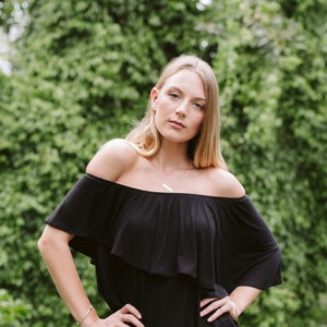 May include: A woman with long blonde hair is wearing a black off-the-shoulder top with a ruffled neckline. She is standing in front of a green leafy background.
