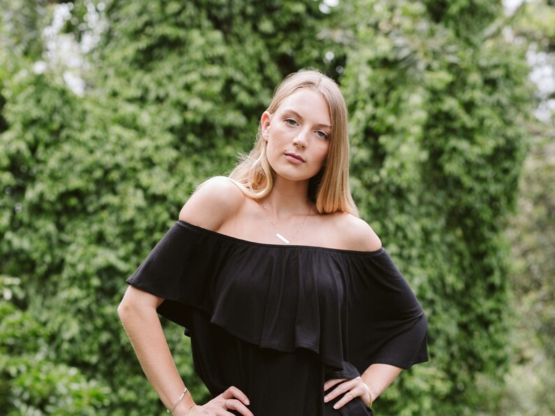 May include: A woman with long blonde hair is wearing a black off-the-shoulder top with a ruffled neckline. She is standing in front of a green leafy background.