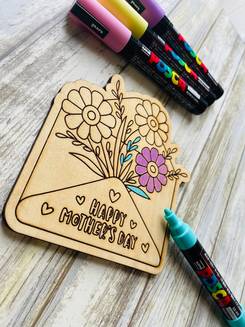 May include: Wooden craft project shaped like an envelope with a bouquet of flowers and the text "Happy Mother's Day" inside. The craft is unfinished and ready to be colored with markers.