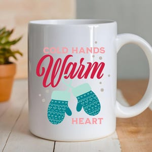 以下が含まれることがあります： ピンクと赤の文字で「COLD HANDS WARM HEART」と書かれた白いセラミックマグ。デザインには、2つのティールミトンが含まれています。小さな鉢植えの植物が背景にあります。