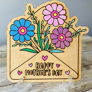 May include: Wooden card with a light brown envelope design. The envelope is filled with a bouquet of flowers in shades of pink, purple, and blue. The card reads "Happy Mother's Day" with pink hearts.