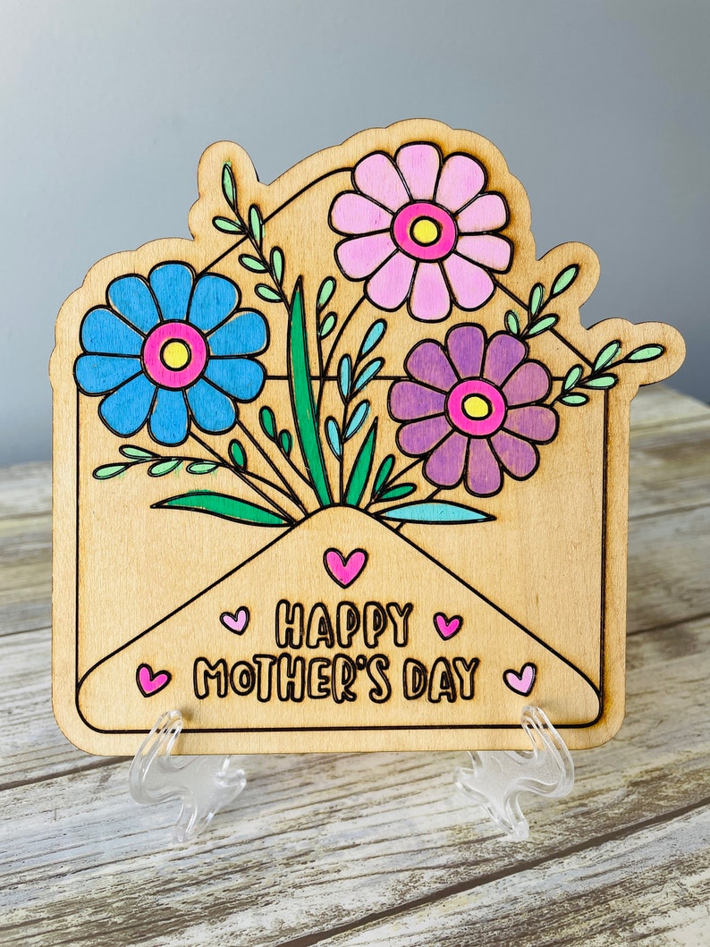May include: Wooden card with a light brown envelope design. The envelope is filled with a bouquet of flowers in shades of pink, purple, and blue. The card reads "Happy Mother's Day" with pink hearts.