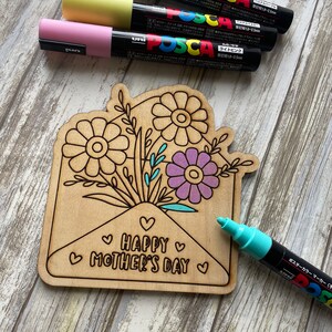 May include: A wooden craft project featuring a flower bouquet inside an envelope with the text "HAPPY MOTHER'S DAY". The project is being colored in with pastel and turquoise Posca paint markers.