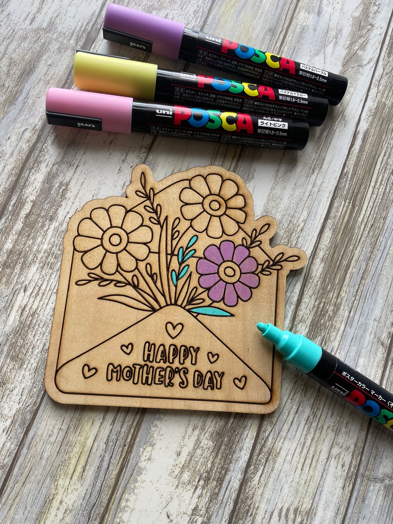 May include: A wooden craft project featuring a flower bouquet inside an envelope with the text "HAPPY MOTHER'S DAY". The project is being colored in with pastel and turquoise Posca paint markers.