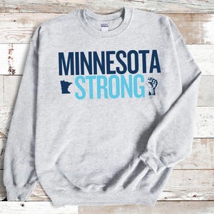 May include: A light gray sweatshirt with the words "MINNESOTA STRONG" in navy and light blue. The design includes a silhouette of the state of Minnesota and a raised fist graphic.
