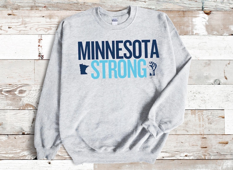 May include: A light gray sweatshirt with the words "MINNESOTA STRONG" in navy and light blue. The design includes a silhouette of the state of Minnesota and a raised fist graphic.