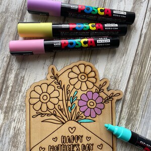 May include: A wooden craft project featuring a floral bouquet inside an envelope with the text "HAPPY MOTHER'S DAY". The project is being colored in with pastel pink, yellow, purple, and turquoise paint markers.