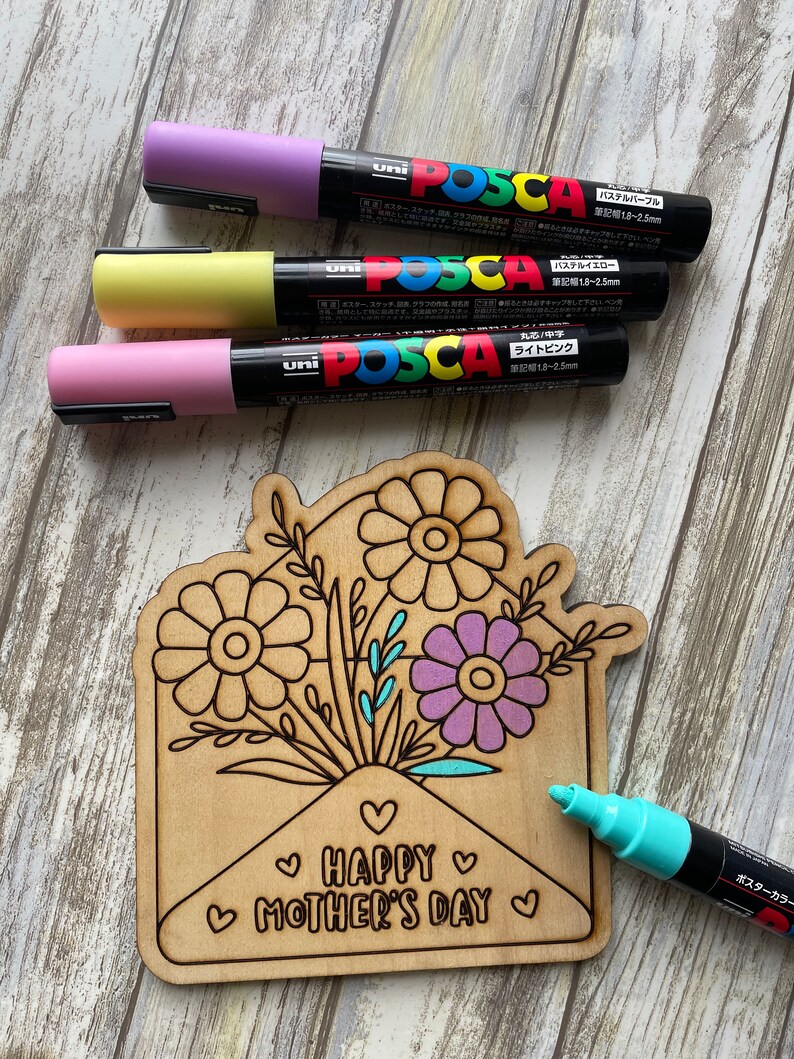 May include: A wooden craft project featuring a floral bouquet inside an envelope with the text "HAPPY MOTHER'S DAY". The project is being colored in with pastel pink, yellow, purple, and turquoise paint markers.
