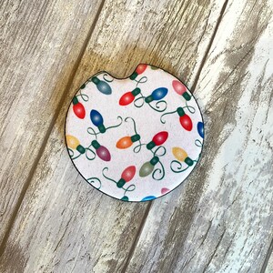 May include: A white round car coaster with a design of colorful Christmas lights strung together. The lights are red, green, yellow, blue, and purple.