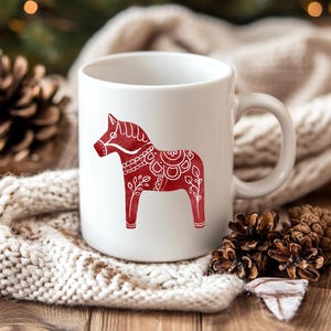 May include: White ceramic mug featuring a red Dala horse design. The horse is decorated with intricate white patterns. The mug has a curved handle and is set against a cozy, neutral-toned background.