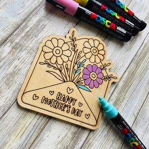 May include: A wooden craft project featuring an envelope with a bouquet of flowers and the text "Happy Mother's Day". The project is surrounded by colorful markers.