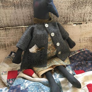 May include: A handcrafted crow doll with a dark blue head and beak, dressed in a grey coat with white buttons. The doll has black limbs and wears a floral dress and shorts. It is positioned on a patchwork quilt, showcasing a rustic aesthetic.
