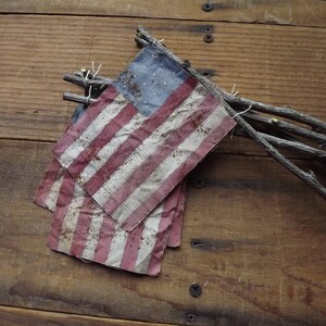 May include: A weathered American flag made of fabric, with red and white stripes and blue stars, is tied to a bundle of twigs.