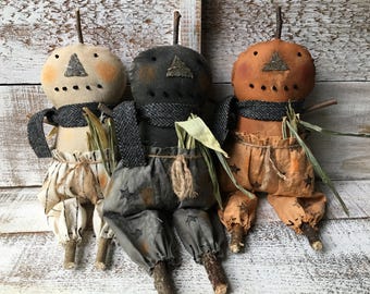 May include: Three fabric dolls with pumpkin-shaped heads, each featuring a twig stem, stitched triangular noses, and simple dot eyes. They wear dark herringbone scarves and pants with star patterns, accented with dried corn husks and twig limbs.
