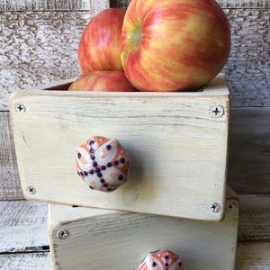 May include: A stack of two small, off-white wooden drawers with decorative ceramic knobs. The drawers are filled with red and yellow apples. The knobs have an orange and blue floral design. The drawers are held together with silver screws.