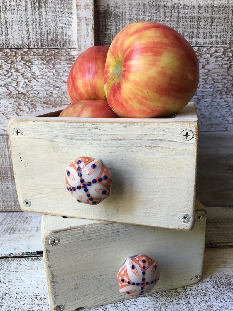 May include: A stack of two small, off-white wooden drawers with decorative ceramic knobs. The drawers are filled with red and yellow apples. The knobs have an orange and blue floral design. The drawers are held together with silver screws.