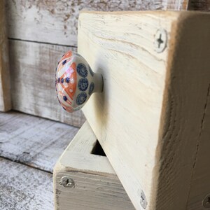May include: Cream-colored wooden block with a decorative ceramic knob. The knob features a pattern of blue, orange, and white floral designs. The block is secured with silver screws, and the wood grain is visible.