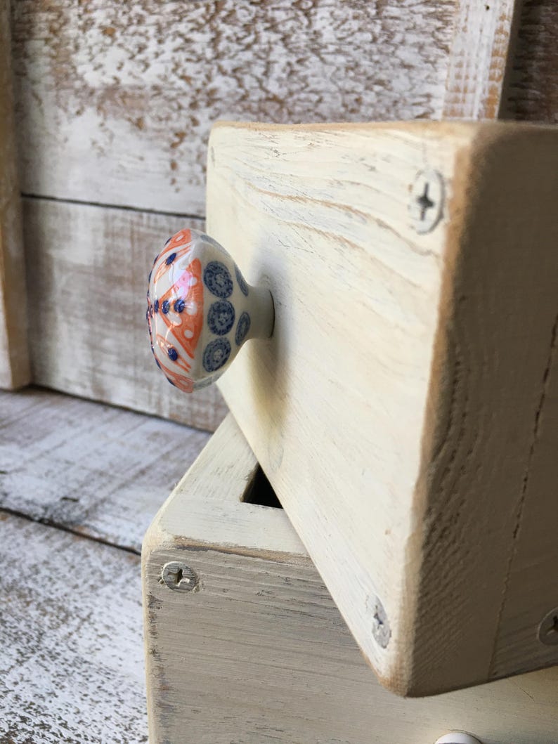 May include: Cream-colored wooden block with a decorative ceramic knob. The knob features a pattern of blue, orange, and white floral designs. The block is secured with silver screws, and the wood grain is visible.
