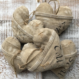 May include: A collection of beige burlap heart-shaped ornaments. Each heart has a stitched design and a number printed in black. Some hearts have twine loops for hanging. The background is a white-painted wooden surface.