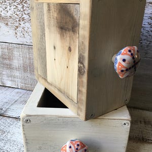 May include: Two wooden boxes, one stacked on top of the other. The top box is natural wood, and the bottom box is painted white. Both have decorative ceramic knobs with orange, blue, and white patterns. The boxes are held together with screws.