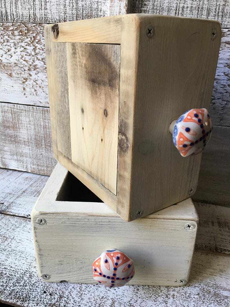 May include: Two wooden boxes, one stacked on top of the other. The top box is natural wood, and the bottom box is painted white. Both have decorative ceramic knobs with orange, blue, and white patterns. The boxes are held together with screws.